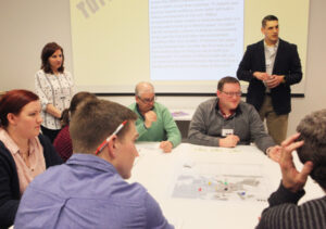 A group of people sit around a table; two people stand in front of a screen presenting